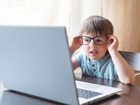 Curious toddler boy explores the laptop and staring on screen through big glasses. Symbol of online education for toddlers or impaired vision from prolonged exposure to screens of electronic devices.