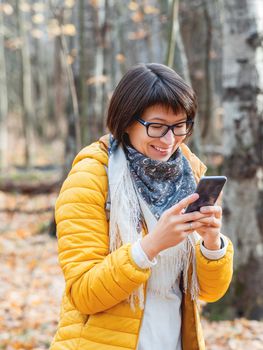 Woman in bright yellow jacket with smartphone. Young girl with wide smile walking in autumn forest and texting someone on smartphone.