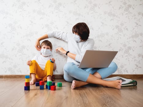 Mother puts protective medical mask on her little son. Family stays at home quarantine because of coronavirus COVID19. Woman remote works with laptop, son plays with toy blocks. Self isolation at home.