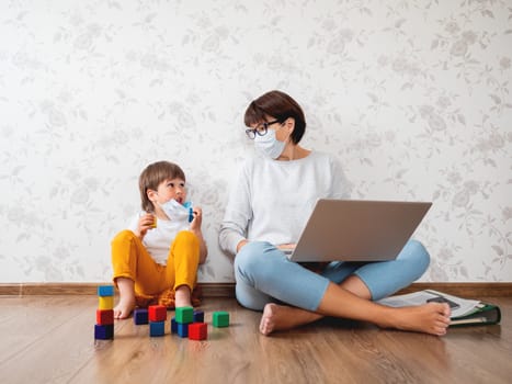 Mother puts protective medical mask on her little son. Family stays at home quarantine because of coronavirus COVID19. Woman remote works with laptop, son plays with toy blocks. Self isolation at home.