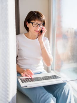 Woman with smartphone remote works from home. She smiles widely on window sill with laptop on knees. Lockdown quarantine because of coronavirus COVID19. Self isolation at home.