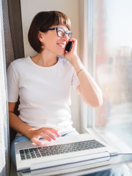 Woman with smartphone remote works from home. She smiles widely on window sill with laptop on knees. Lockdown quarantine because of coronavirus COVID19. Self isolation at home.