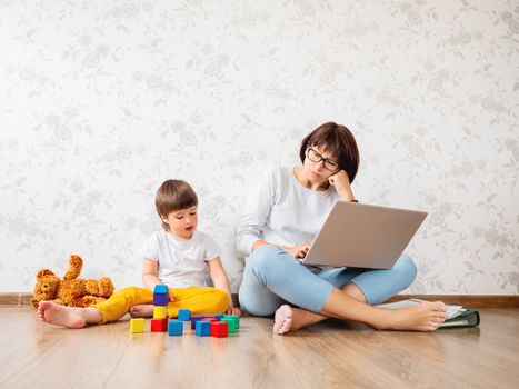 Mom and son sit at home quarantine because of coronavirus COVID19. Mother remote works with laptop, son plays with toy blocks. Self isolation at home.