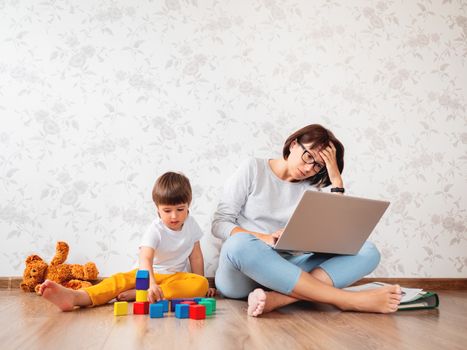 Mom and son sit at home quarantine because of coronavirus COVID19. Mother remote works with laptop, son plays with toy blocks. Self isolation at home.