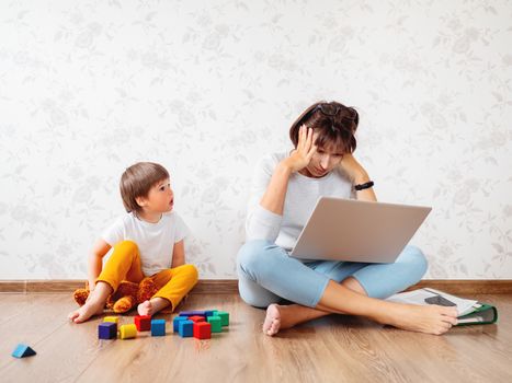 Mom and son sit at home quarantine because of coronavirus COVID19. Mother remote works with laptop, son plays with toy blocks. Self isolation at home.