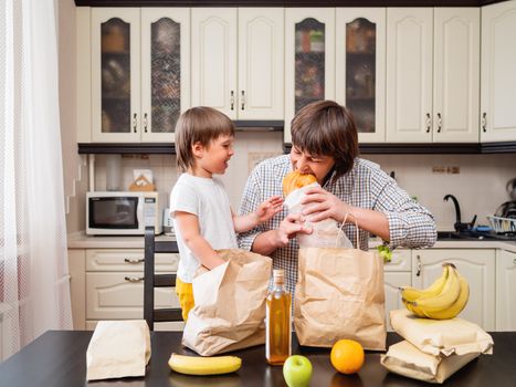 Family sorts out purchases in the kitchen. Father and son tastes products in bags made of craft paper. Food delivery in conditions of quarantine because of coronavirus COVID19.