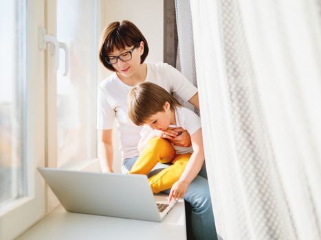 Woman remote works from home. She sits on window sill with laptop and cute toddler boy on her knees. Lockdown quarantine because of coronavirus COVID19. Self isolation at home.