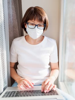 Woman in medical mask remote works from home. She sits on window sill with laptop on knees. Lockdown quarantine because of coronavirus COVID19. Self isolation at home.