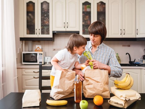 Family sorts out purchases in the kitchen. Father and son tastes products in bags made of craft paper. Food delivery in conditions of quarantine because of coronavirus COVID19.