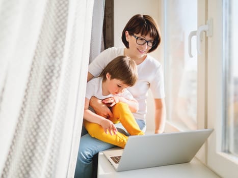 Woman remote works from home. She sits on window sill with laptop and cute toddler boy on her knees. Lockdown quarantine because of coronavirus COVID19. Self isolation at home.