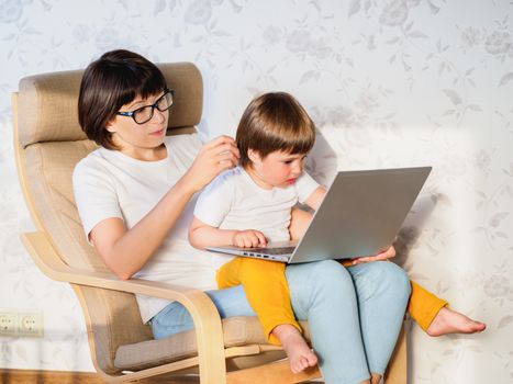 Mother and her toddler boy sit together on chair with laptop. Woman tries to remote work, but kid is asking for game or cartoons. quarantine lockdown because of coronavirus COVID-19.