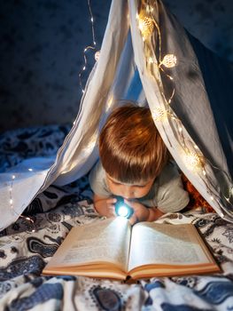 Little boy reads with pocket light. Toddler plays in tent made of linen sheet on bed. Cozy evening with favorite book.