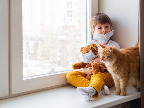 Toddler boy sits with teddy bear, both in medical masks. Kid with cute ginger cat. Fluffy pet and child on home quarantine because of coronavirus COVID-19.