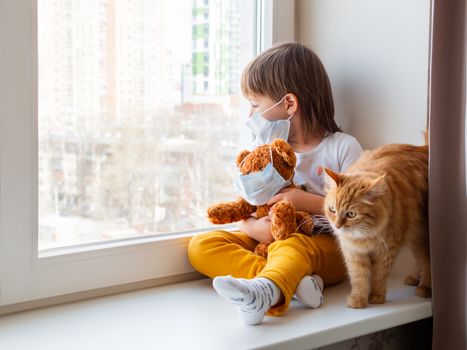 Toddler boy sits with teddy bear, both in medical masks. Kid with cute ginger cat. Fluffy pet and child on home quarantine because of coronavirus COVID-19.