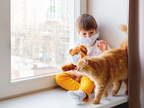 Toddler boy sits with teddy bear, both in medical masks. Kid with cute ginger cat. Fluffy pet and child on home quarantine because of coronavirus COVID-19.