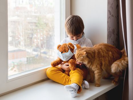 Toddler boy sits with teddy bear, both in medical masks. Kid with cute ginger cat. Fluffy pet and child on home quarantine because of coronavirus COVID-19.