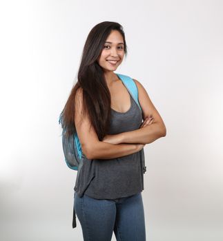 A young female student in casual clothes crosses her arms as she smiles and holds her backpack.