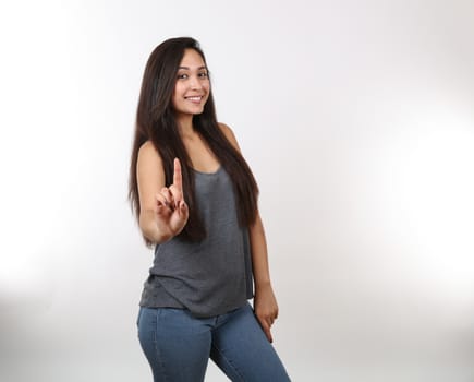 A young attractive girl smiles and points her finger up.  Wearing blue jeans and a grey tank top.