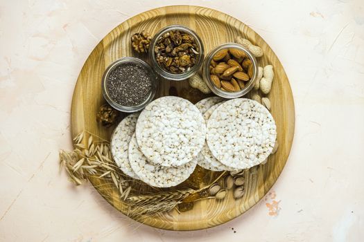 American puffed rice cakes. Healthy snacks with ears of wheat on round cutting wooden board on light pink concrete surface, top view, flat lay, copy space for you text	