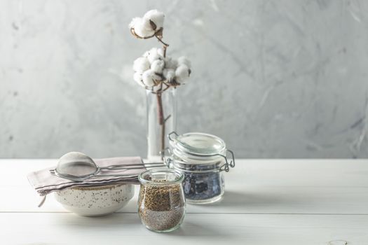 Coffee beans and instant coffee in glass jars, cotton flowers and kitchenware on white wooden table with grey concrete wall at background. Details of still life in the home interior. Cosy concept.