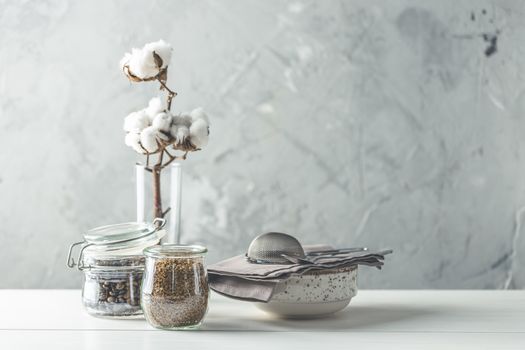 Coffee beans and instant coffee in glass jars, cotton flowers and kitchenware on white wooden table with grey concrete wall at background. Details of still life in the home interior. Cosy concept.