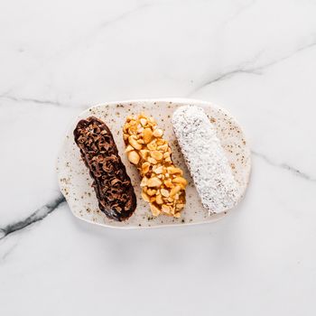 Set of three homemade eclairs on marble background. Top view of delicious healthy profitroles with different decor ingredients - chocolate, peanut and sherdded coconut. Square
