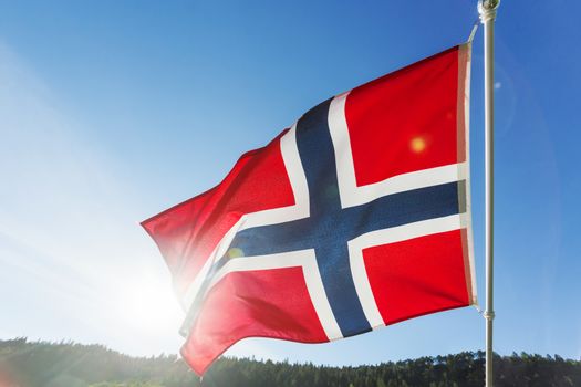 Flag of Norway waving in the wind against summer forest landscape in sunny day.