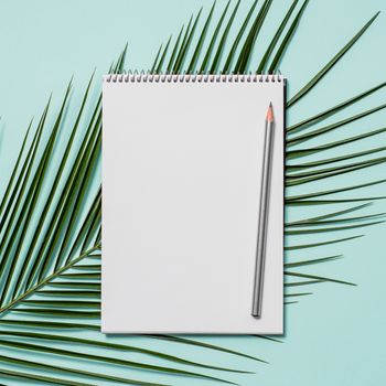 Blank paper notebook with pencil. Empty paper sketchbook with pencil and tropical leaf on blue background. Top view or flat lay. Copy space for text or design.