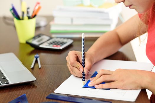 Young schoolgirl is doing a homework with math and geometry next to a laptop, calculator and books on her desk in her room