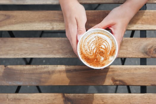 Woman holding a paper cup with coffee. Coffee to go. Tasty hot beverage on wooden table in sunny day. Outdoors meal. Flat lay, top view.