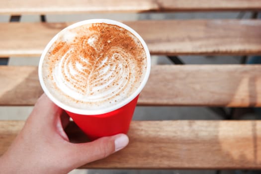 Woman holding a red paper cup with coffee. Coffee to go. Tasty hot beverage on wooden table in sunny day. Outdoors meal.