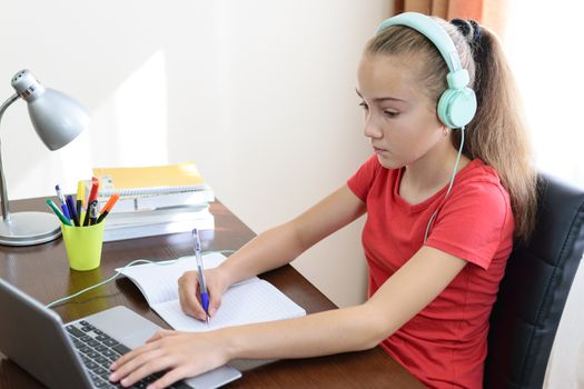 Young schoolgirl with headphones is doing a homework or learning foreign language on the laptop in her room.