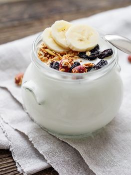 Natural homemade yogurt in a glass jar. Healthy food for breakfast with muesli. Jar with granola and banana slices on linen tablecloth on on wooden table.