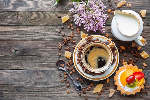 Rustic wooden background with cup of coffee, milk, fruit tart and lilac flowers. White vintage dinnerware and spoon. Breakfast at summer morning. Top view, place for text.