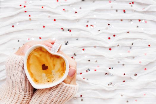 Woman holds a cup of hot coffee with cinnamon heart. Winter and Valentine's Day fabric background with sparkling silver and red confetti.