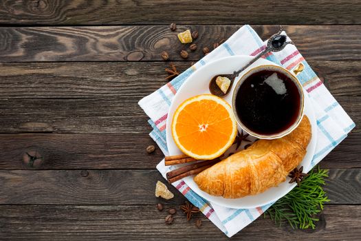 Continental breakfast - cup of hot coffee, croissant and orange. Tasty food on plaid napkin. Rustic wooden background. Top view, place for text.
