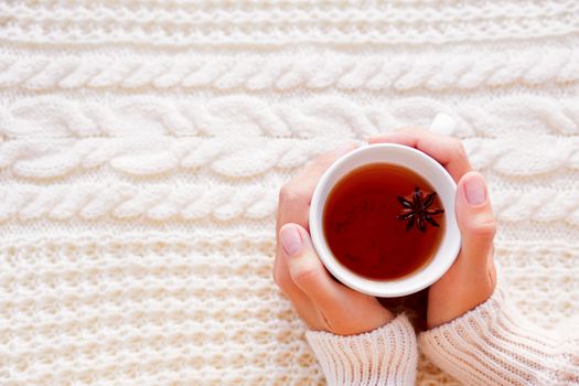 Women holds a cup of hot tea with anise star. Beautiful fabric background with knitted pattern. Cozy morning at home.