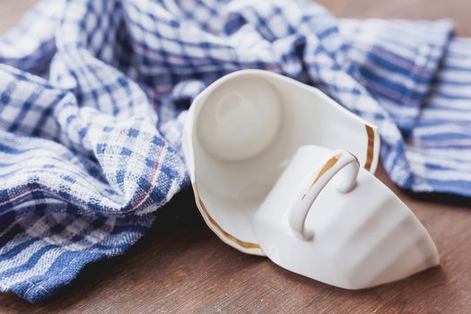 Broken white cup on wooden background with plaid blue towel. Damaged mug with golden decoration.
