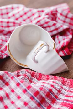 Broken white cup on wooden background with plaid red towel. Damaged mug with golden decoration.