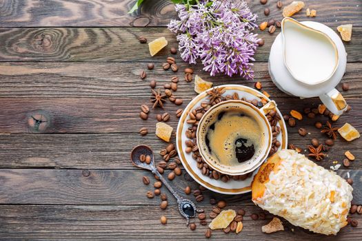 Rustic wooden background with cup of coffee, milk, bun with marzipan and lilac flowers. White vintage dinnerware and spoon. Breakfast at summer morning. Top view, place for text.