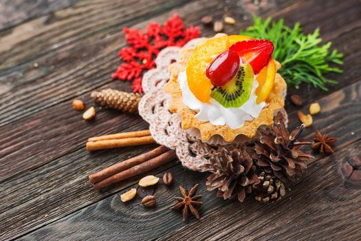 Winter holiday rustic background with fruit tart and spices - cinnamon, anise, peanuts. Christmas and New Year decorations - red snowflake, thuja and pine cones. Tasty dessert with strawberry, kiwi, orange, peach and whipped cream.