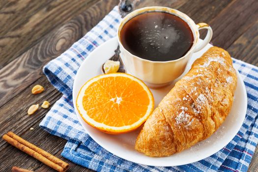 Continental breakfast - cup of hot coffee, croissant and orange. Tasty food on plaid blue napkin, rustic wooden background.