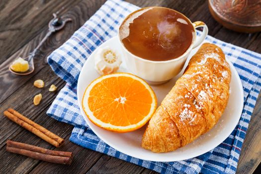 Continental breakfast - cup of hot coffee, croissant and orange. Tasty food on plaid blue napkin, rustic wooden background.