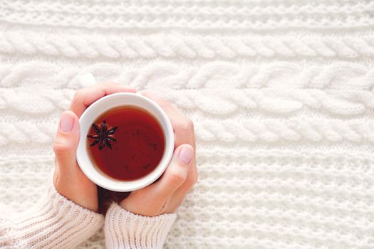 Women holds a cup of hot tea with anise star. Beautiful fabric background with knitted pattern. Cozy morning at home.