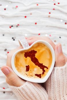 Woman holds a cup of hot coffee with cinnamon silhouette of Italy. Winter wanderlust and Valentine's Day fabric background with sparkling silver and red confetti.