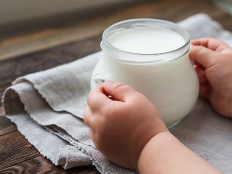 Natural homemade yogurt in a glass jar. Healthy food for breakfast in child's hands. Kid holding jar with sour cream on linen tablecloth on on wooden table.