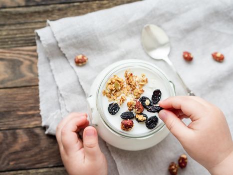 Natural homemade yogurt in a glass jar. Healthy food for breakfast in child's hands. Kid holding jar with muesli on linen tablecloth on on wooden table.