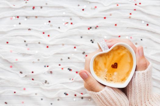 Woman holds a cup of hot coffee with cinnamon heart. Winter and Valentine's Day fabric background with sparkling silver and red confetti.