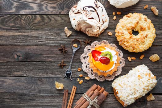 Rustic wooden background with different pastries - chocolate meringue, fruit tart, marzipan bun. Different spices - cinnamon, walnuts, peanuts, ginger. Top view of tasty desserts. Place for text.