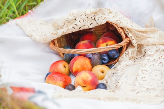 Fruit basket with peaches, plums and pears lying on the lace fabric. Picnic outdoor.